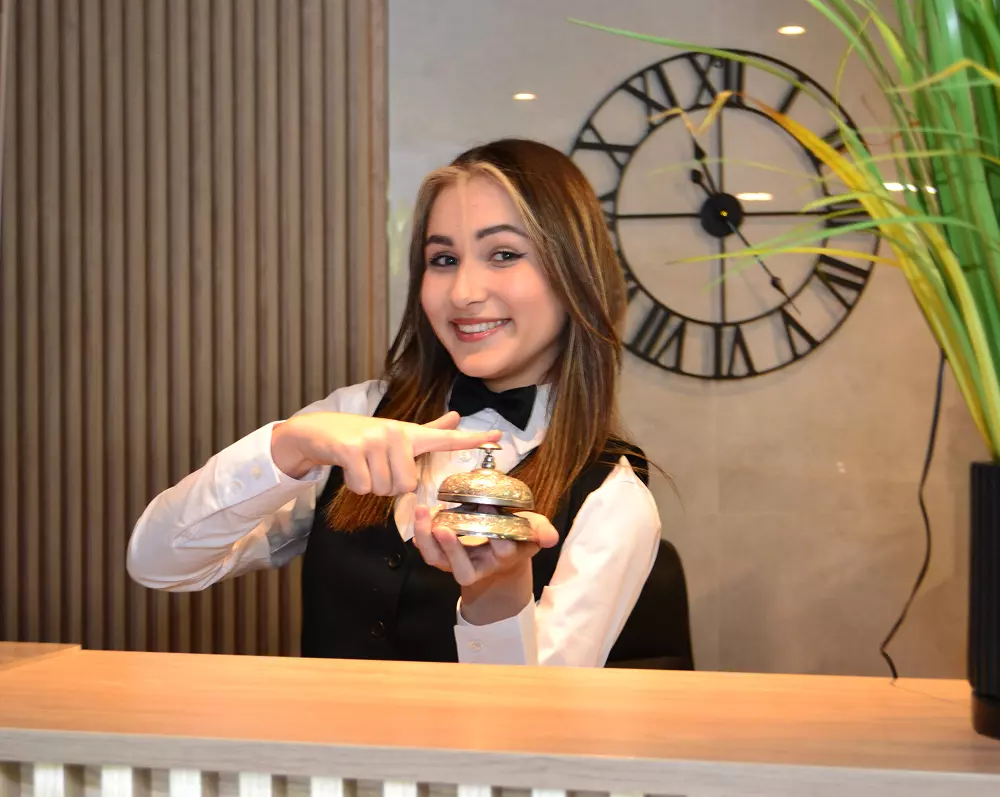 Uczennica kierunku hotelarstwo ubrana w uniform stoi na recepcji i pokazuje dzwonek recepcyjny